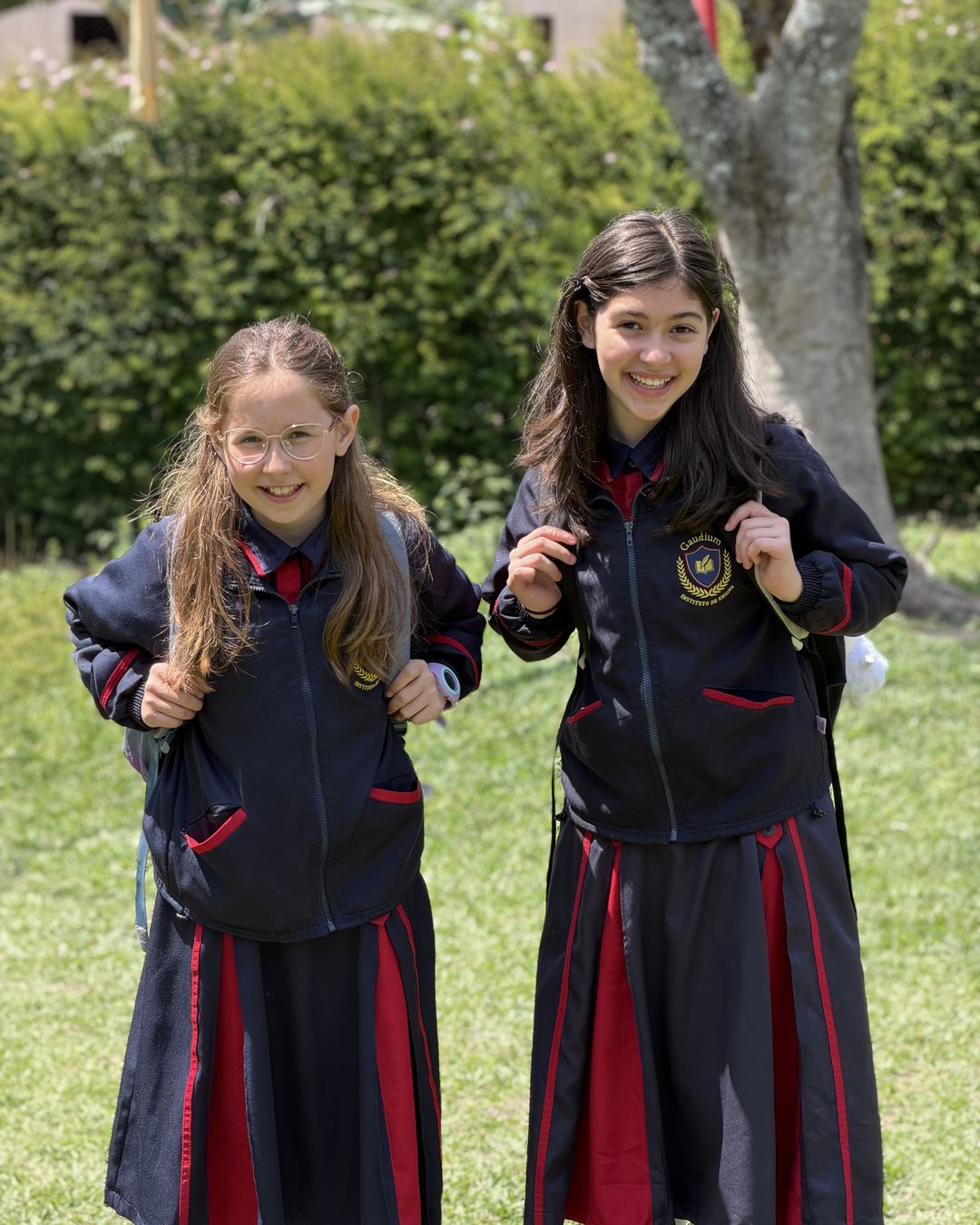 Duas alunas do Colégio Gaudium sorrindo de mochila no jardim da escola